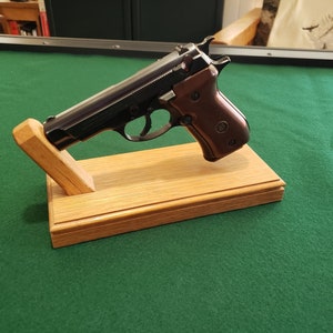 Könnte beinhalten: Eine schwarze Pistole mit braunem Griff wird auf einem Holzständer präsentiert. Der Ständer ist aus hellem braunem Holz gefertigt und hat ein schlichtes Design.