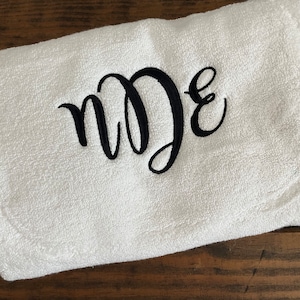 May include: A white rectangular terry cloth towel featuring black embroidered script lettering in the center. The embroidery displays the letters n, D, and E in a stylized, interconnected cursive font, creating a monogram design on the fabric.
