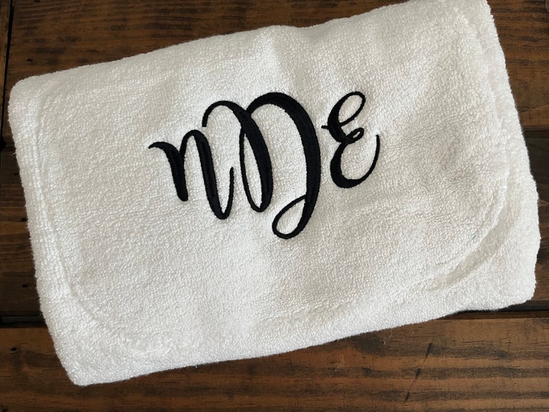 May include: A white rectangular terry cloth towel featuring black embroidered script lettering in the center. The embroidery displays the letters n, D, and E in a stylized, interconnected cursive font, creating a monogram design on the fabric.