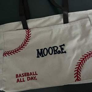 May include: Cream-colored tote bag with baseball-themed embroidery. Features red stitching mimicking a baseball's seams, the name "MOORE" in navy blue, and the phrase "BASEBALL ALL DAY." Includes brown leather handles and side pockets.