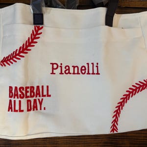 May include: A white canvas tote bag with red baseball stitching and the words "Pianelli" and "BASEBALL ALL DAY" embroidered in red. The bag has black handles and side pockets.
