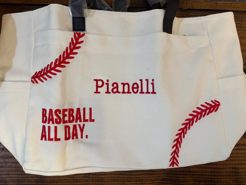 May include: A white canvas tote bag with red baseball stitching and the words "Pianelli" and "BASEBALL ALL DAY" embroidered in red. The bag has black handles and side pockets.