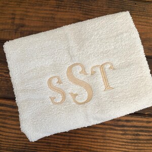 May include: A folded white terry cloth towel featuring an embroidered monogram in a light tan thread. The monogram consists of the letters S, S, and T in a decorative serif font, centered on the fabric surface of the rectangular towel.