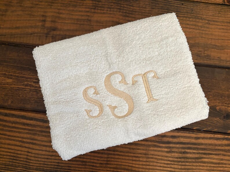 May include: A folded white terry cloth towel featuring an embroidered monogram in a light tan thread. The monogram consists of the letters S, S, and T in a decorative serif font, centered on the fabric surface of the rectangular towel.