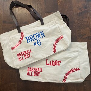 May include: Two off-white canvas tote bags with brown leather handles. One bag has "BROWN #6" in blue and "BASEBALL ALL DAY" in red. The other bag says "Liner" in red and "BASEBALL ALL DAY" in red. Both bags have red baseball stitching.