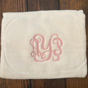 May include: A folded off-white terry cloth towel featuring a decorative pink embroidered monogram in the center. The embroidery consists of three intertwined cursive letters, showcasing a detailed and elegant script design on the soft fabric.