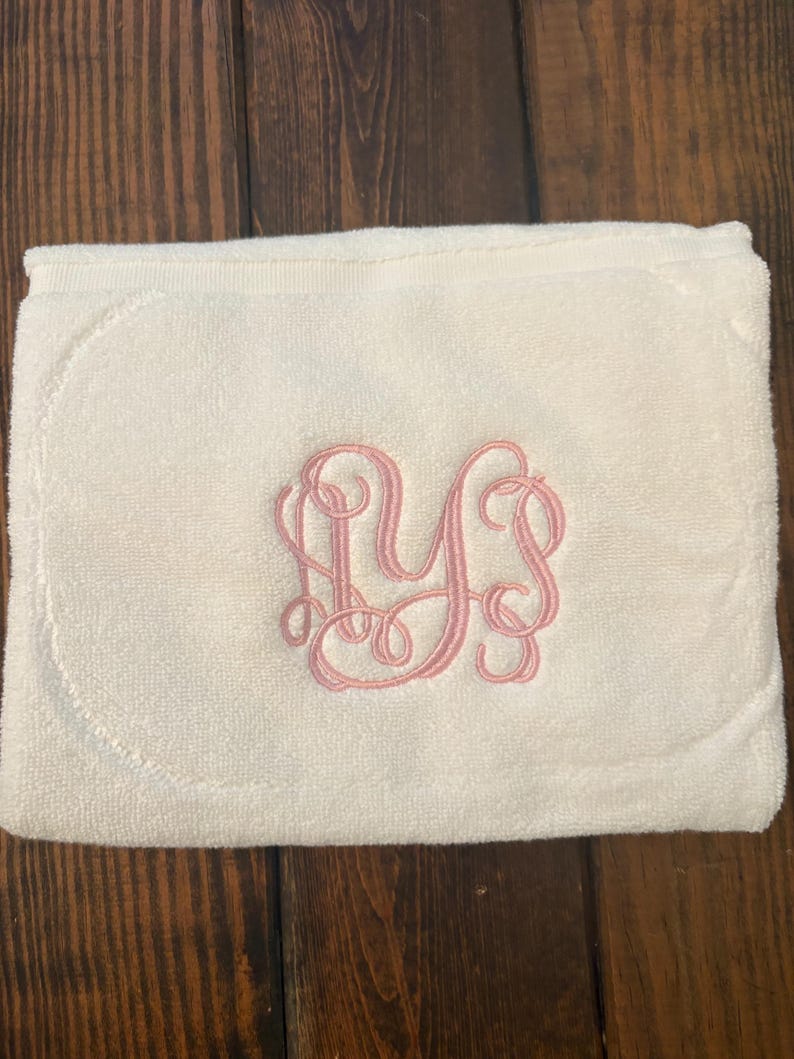 May include: A folded off-white terry cloth towel featuring a decorative pink embroidered monogram in the center. The embroidery consists of three intertwined cursive letters, showcasing a detailed and elegant script design on the soft fabric.