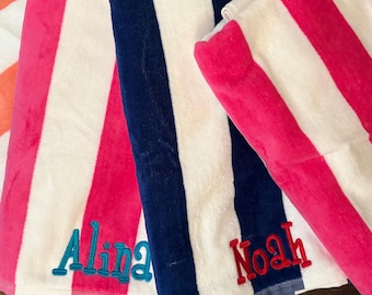 May include: A collection of four striped beach towels featuring vertical bands in orange, hot pink, and navy blue alternating with white. Each towel is personalized with embroidered names in contrasting thread colors: Sullivan, Alina, Noah, and Cora.