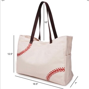 May include: Cream-colored baseball-themed tote bag with brown leather handles. The bag features red stitching details mimicking a baseball design. Dimensions: 16.5" wide, 12.5" high, 7" deep, with 10" handle drop.
