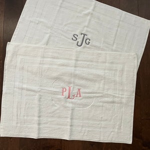May include: Two rectangular white bath mats featuring a textured border design. The top mat displays the embroidered gray monogram SJG, while the bottom mat features the embroidered pink monogram PLA centered within the rectangular frame.