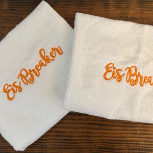 May include: Two folded white towels featuring the text Eis Breaker embroidered in bright orange thread. The towels have a soft, textured surface and are positioned side by side, displaying the same cursive lettering on each piece of fabric.