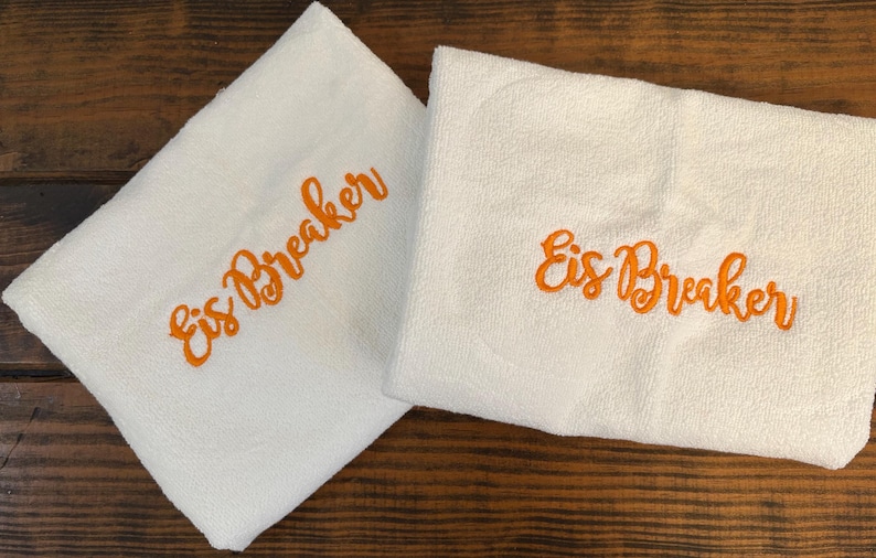 May include: Two folded white towels featuring the text Eis Breaker embroidered in bright orange thread. The towels have a soft, textured surface and are positioned side by side, displaying the same cursive lettering on each piece of fabric.