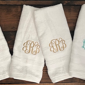 May include: Six white terry cloth towels arranged in pairs, each featuring a decorative embroidered monogram. The first pair has pink lettering, the second pair has gold lettering, and the third pair has light teal lettering, all in a script font.