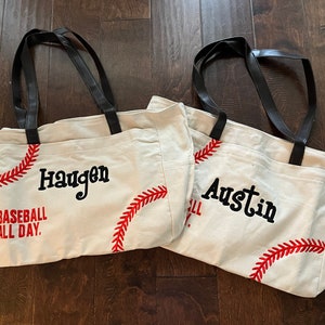 May include: Two canvas tote bags with black handles and baseball stitching. One bag has the name "Haugen" and the text "Baseball All Day." The other bag has the name "Austin" and baseball stitching.