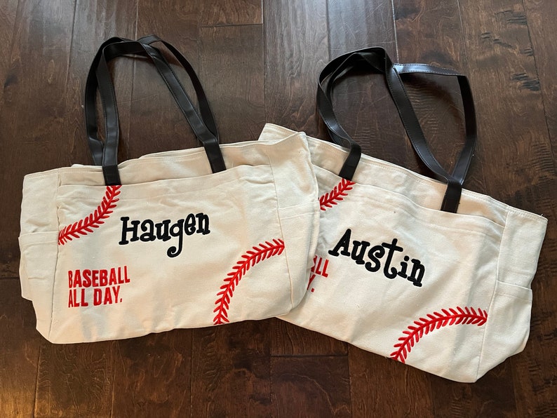 May include: Two canvas tote bags with black handles and baseball stitching. One bag has the name "Haugen" and the text "Baseball All Day." The other bag has the name "Austin" and baseball stitching.