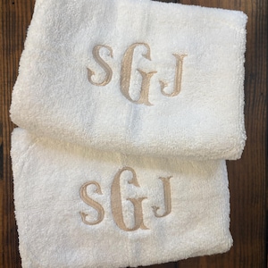 May include: Two folded white terry cloth towels featuring an embroidered monogram in a light tan thread. Each towel displays the letters SGJ in a decorative serif font, with the central letter G being larger than the S and J on either side.