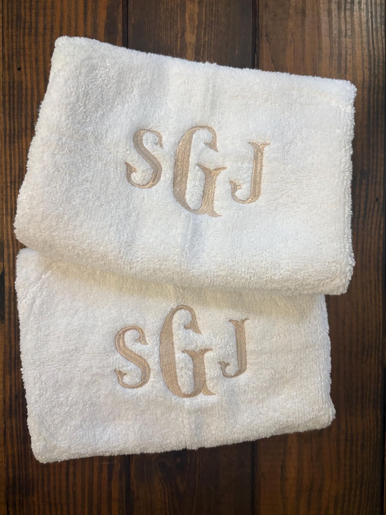 May include: Two folded white terry cloth towels featuring an embroidered monogram in a light tan thread. Each towel displays the letters SGJ in a decorative serif font, with the central letter G being larger than the S and J on either side.