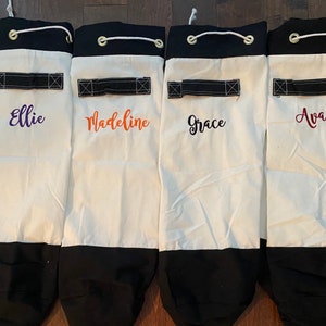 May include: Four white canvas drawstring bags with black trim and black straps. Each bag has a different name embroidered on the front: Ellie, Madeline, Grace, and Ava.
