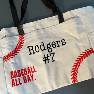 May include: Cream-colored tote bag with black handles and side pockets. The bag features red baseball stitching and the text "Rodgers #7" and "Baseball All Day" in black and red embroidery. The bag is made of a sturdy fabric.