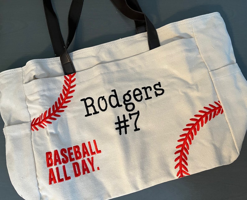 May include: Cream-colored tote bag with black handles and side pockets. The bag features red baseball stitching and the text "Rodgers #7" and "Baseball All Day" in black and red embroidery. The bag is made of a sturdy fabric.