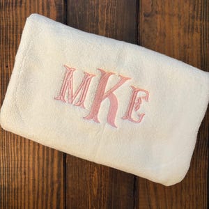 May include: A folded off-white towel featuring an embroidered monogram in a soft pink thread. The letters MKE are stitched in a decorative serif font centered on the fabric. The towel has a plush, textured surface and is rectangular in shape.