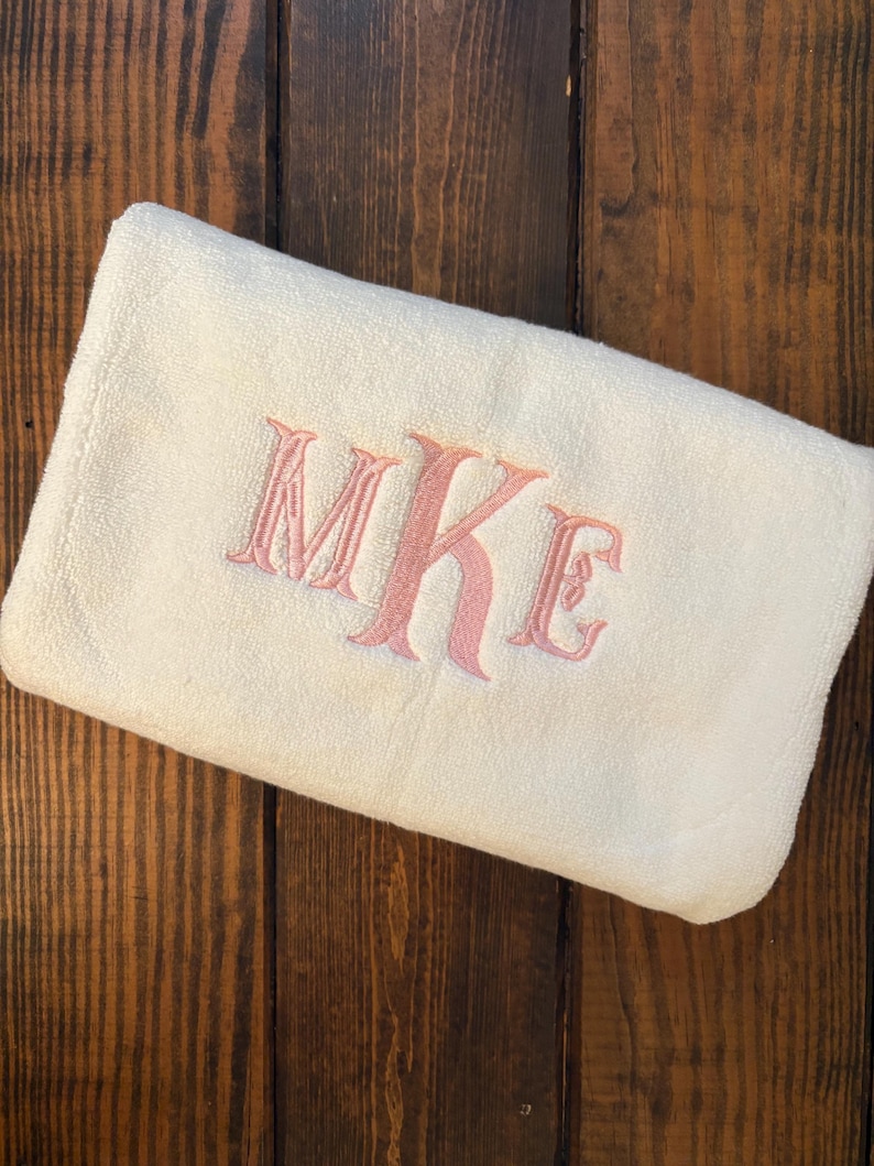 May include: A folded off-white towel featuring an embroidered monogram in a soft pink thread. The letters MKE are stitched in a decorative serif font centered on the fabric. The towel has a plush, textured surface and is rectangular in shape.