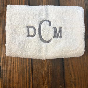 May include: A white rectangular terry cloth towel featuring a centered embroidered monogram in grey thread. The monogram consists of the letters D and M in a standard serif font, flanking a larger, central letter C in a script font.