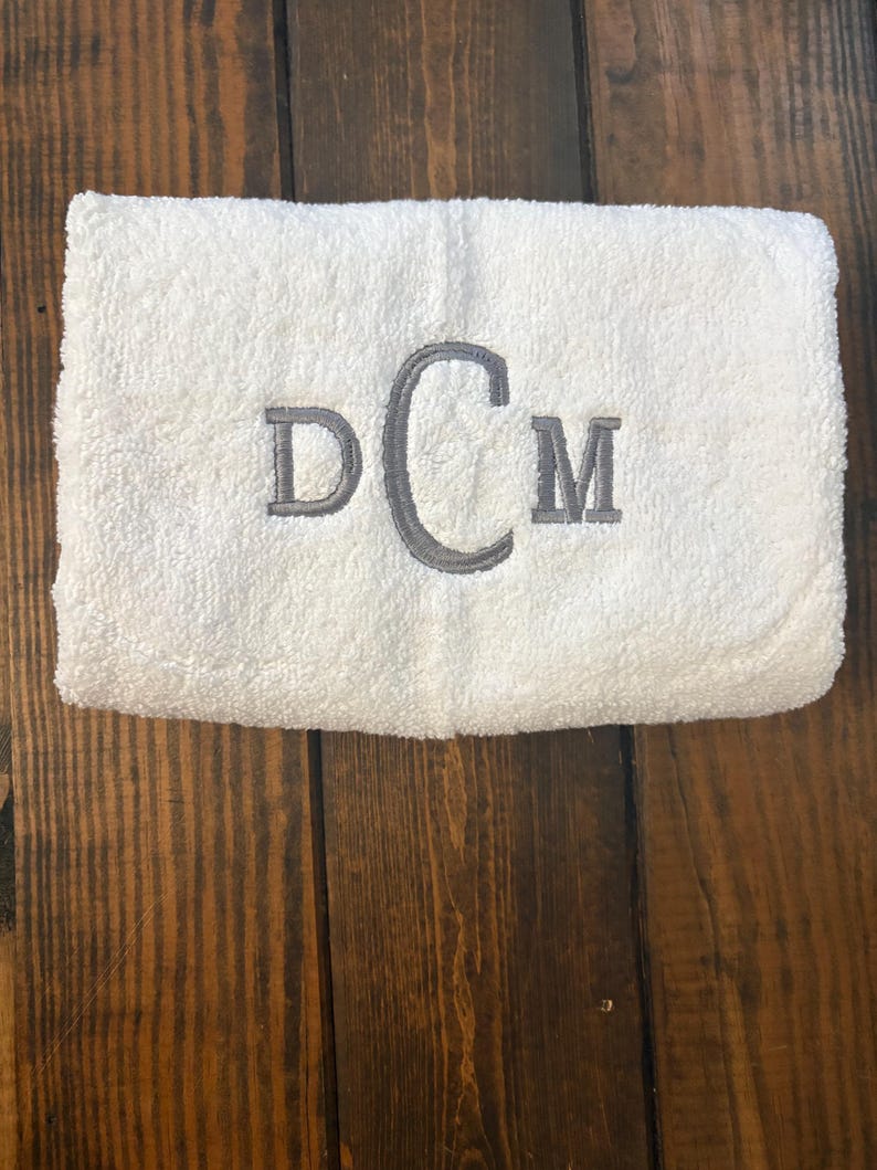 May include: A white rectangular terry cloth towel featuring a centered embroidered monogram in grey thread. The monogram consists of the letters D and M in a standard serif font, flanking a larger, central letter C in a script font.