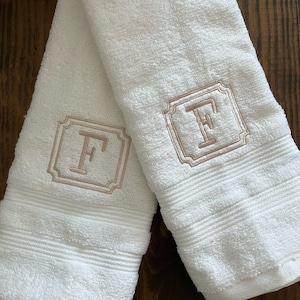 May include: Two white terry cloth hand towels featuring a centered, embroidered monogram of the letter F in a light tan thread. The letter is enclosed within a decorative square frame with notched corners, and each towel has horizontal ribbed bands.