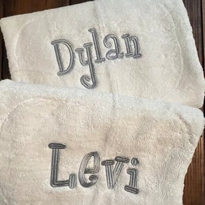 May include: Two white terry cloth towels featuring embroidered names in a metallic silver thread. The top towel displays the name Dylan in a cursive font, while the bottom towel features the name Levi in a matching silver embroidered script.