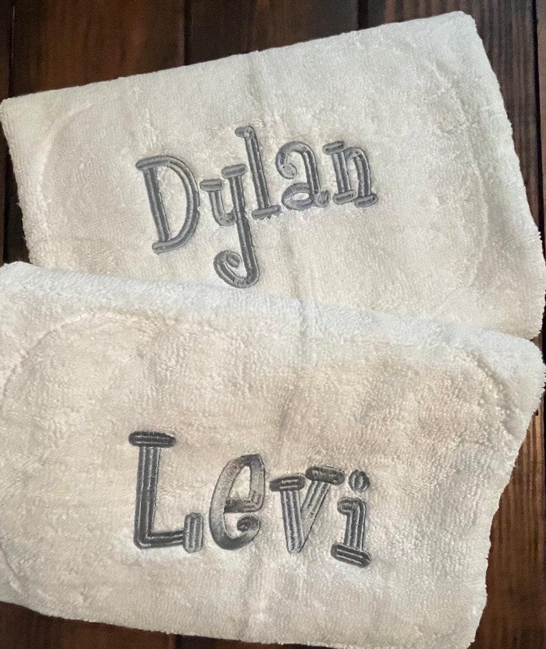 May include: Two white terry cloth towels featuring embroidered names in a metallic silver thread. The top towel displays the name Dylan in a cursive font, while the bottom towel features the name Levi in a matching silver embroidered script.