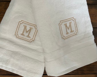 May include: Two white terry cloth towels featuring a centered, embroidered beige letter M enclosed within a decorative square frame. Each towel has a textured border with horizontal ribbed lines near the bottom edge of the fabric.