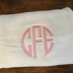 May include: A folded white towel featuring a circular monogram embroidery in light pink thread. The monogram consists of the letters C, F, and E, with the central letter F being larger than the flanking letters C and E, all enclosed in a circle.