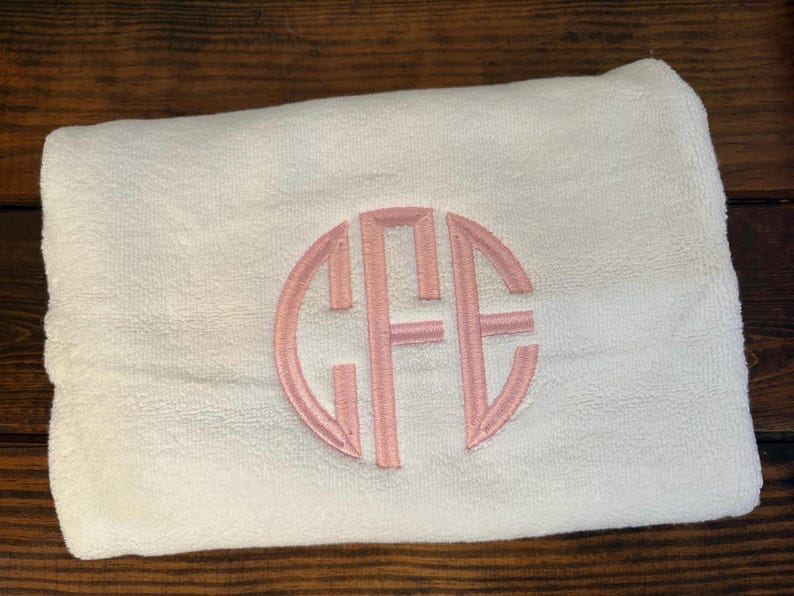 May include: A folded white towel featuring a circular monogram embroidery in light pink thread. The monogram consists of the letters C, F, and E, with the central letter F being larger than the flanking letters C and E, all enclosed in a circle.