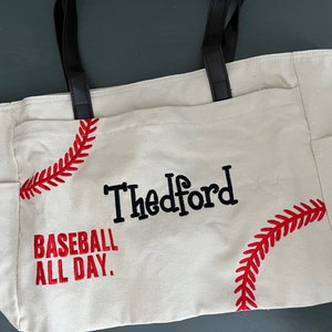 May include: Cream-colored tote bag with black handles and side pockets. The bag features red baseball stitching designs and the text "Thedford BASEBALL ALL DAY" in black and red. A practical and stylish accessory.