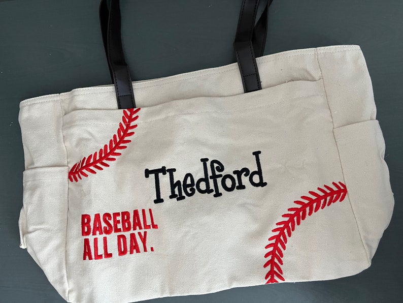 May include: Cream-colored tote bag with black handles and side pockets. The bag features red baseball stitching designs and the text "Thedford BASEBALL ALL DAY" in black and red. A practical and stylish accessory.