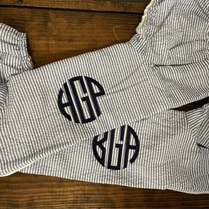 May include: Two white and blue striped fabric pieces with a navy blue monogram on each. The monogram on the top piece is "HOP" and the monogram on the bottom piece is "KUA".
