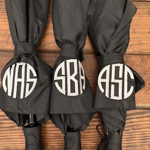 May include: Three black umbrellas with white monogrammed initials. Each umbrella has a different set of initials: NAS, SRA, and ASC. The umbrellas are closed and have black handles.