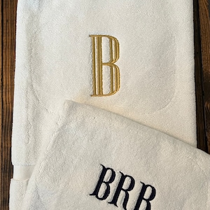 May include: Two white cotton towels featuring embroidered monograms. The larger towel displays a single gold letter B in a serif font, while the smaller towel features the letters BRB embroidered in a dark navy blue serif font.