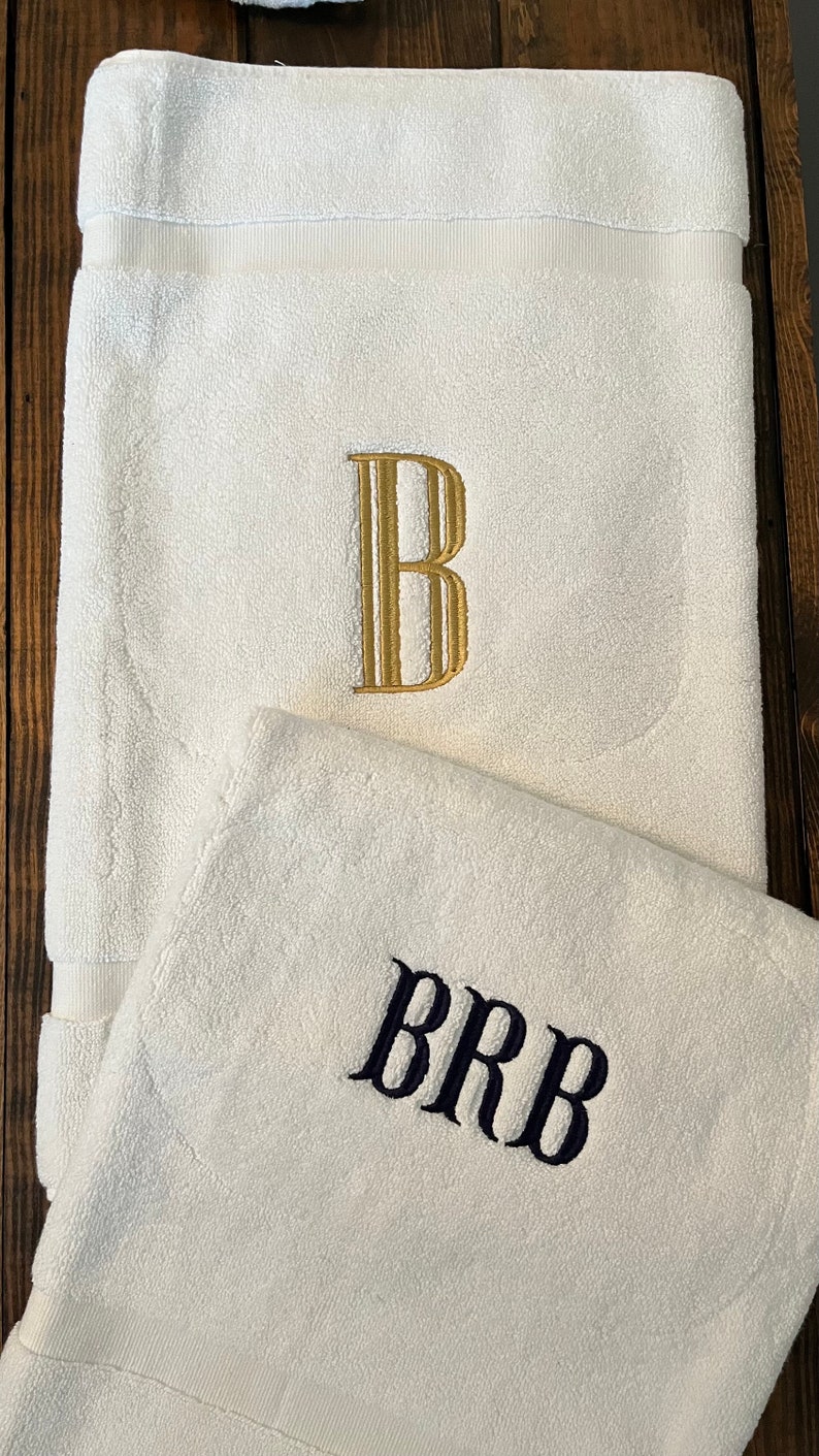 May include: Two white cotton towels featuring embroidered monograms. The larger towel displays a single gold letter B in a serif font, while the smaller towel features the letters BRB embroidered in a dark navy blue serif font.