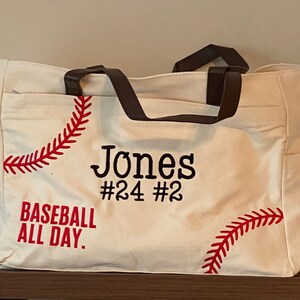 May include: Cream-colored baseball tote bag with brown handles and side pockets. The bag features red baseball stitching and the name "Jones" with the numbers "#24 #2" in black. The text "BASEBALL ALL DAY" is also in red.