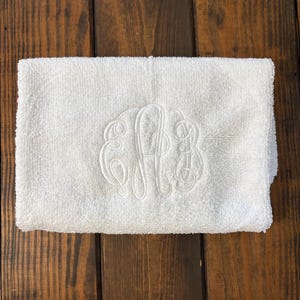 May include: A white rectangular hand towel featuring a centered, tone-on-tone embroidered monogram with three intertwined script letters. The towel has a soft, textured terry cloth surface and is folded neatly into a compact, rectangular shape.