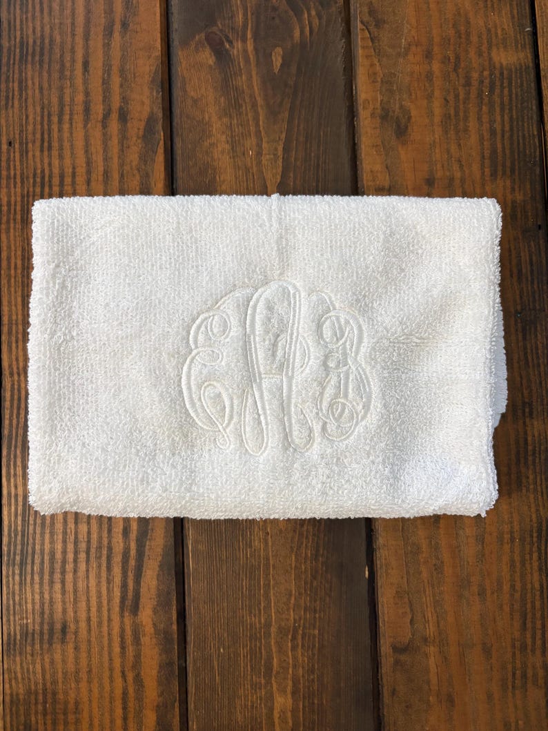May include: A white rectangular hand towel featuring a centered, tone-on-tone embroidered monogram with three intertwined script letters. The towel has a soft, textured terry cloth surface and is folded neatly into a compact, rectangular shape.