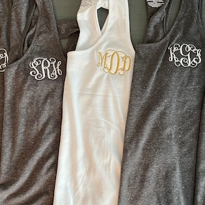 May include: Five tank tops with different monograms embroidered on the front. The tank tops are in grey and white.