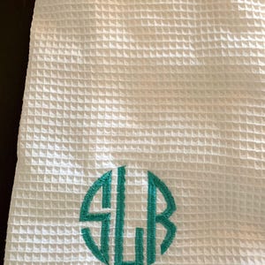 May include: White waffle-weave robe with a teal monogram. The robe has a textured, grid-like pattern and a folded collar. The monogram is a circular design with the letters SLB.