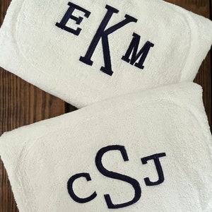 May include: Two white rectangular terry cloth towels featuring dark navy blue embroidery. The top towel displays the monogram EKM in a serif font, while the bottom towel features the monogram cSJ in a rounded, cursive-style script font.
