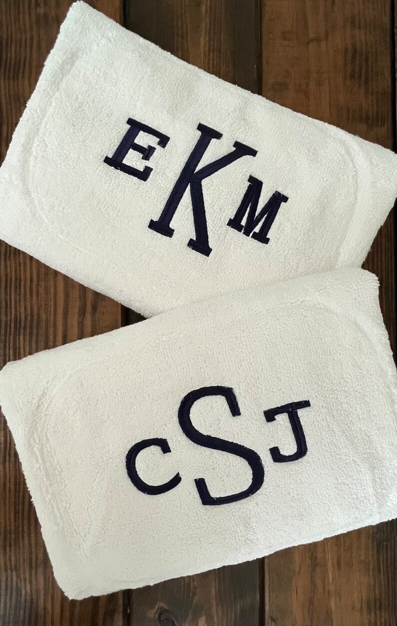 May include: Two white rectangular terry cloth towels featuring dark navy blue embroidery. The top towel displays the monogram EKM in a serif font, while the bottom towel features the monogram cSJ in a rounded, cursive-style script font.