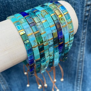 May include: A stack of colorful beaded bracelets in shades of blue, green, and gold. The bracelets are made of small, square beads and have a tassel at the end.