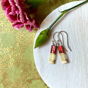 May include: A pair of dangle earrings with red, gold, and white glass beads. The earrings are on a white surface with a green background.