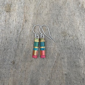 May include: A pair of dangle earrings with a multi-colored, layered design. The earrings are made of small, rectangular beads in shades of green, blue, gold, and red. The beads are strung on a silver wire and have a silver hook.